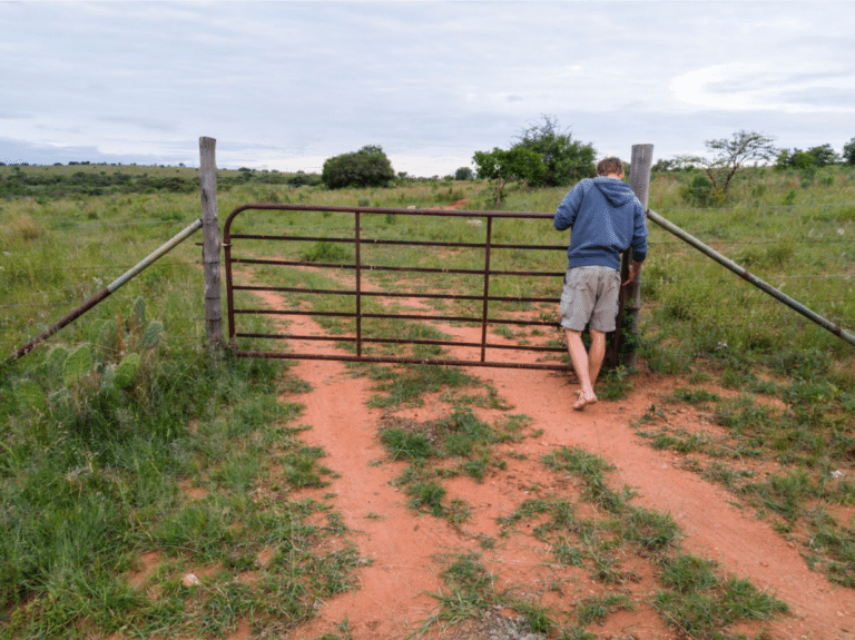 Blogs 1 A person in a hoodie opening a rusty farm gate on a dirt path surrounded by grass.