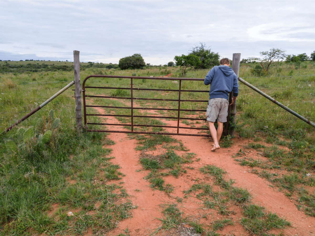 A person in a hoodie opening a rusty farm gate on a dirt path surrounded by grass.